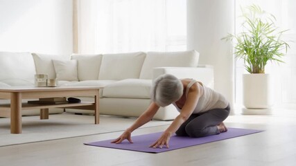 A senior woman practices yoga on a mat in a bright, modern living room. The room has natural light and a calming atmosphere, featuring a sofa and a potted plant Stock Video - Powered by Adobe