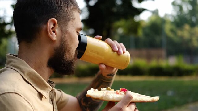 Bearded man enjoys a slice of pizza and drinks tea from a yellow thermos mug while relaxing in a park on a sunny afternoon. Casual outdoor lifestyle. 4k horizontal footage