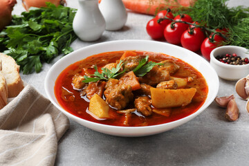 Delicious cooked stew in bowl with fresh ingredients and bread on grey table, closeup