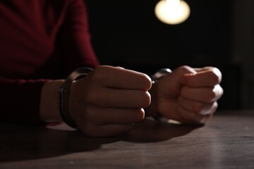 Woman in metal handcuffs at wooden table indoors, closeup