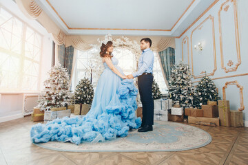 a young pregnant girl in a lush festive blue dress and a young man in a blue shirt. man holding woman's hand. expecting a child. Christmas mood. married couple. family photo session. family photo