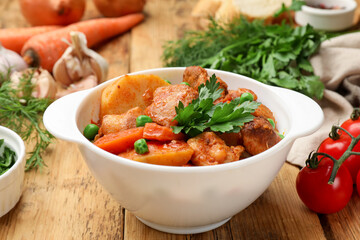 Delicious cooked stew in bowl and ingredients on wooden table, closeup