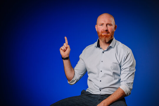 Bearded man with short hair in a light shirt is sitting against a blue background, raising his finger to indicate a concept or idea, showcasing a thoughtful expression