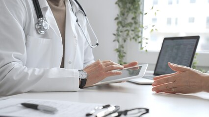 Fototapeta premium Male doctor in lab coat and stethoscope listening to patient while using tablet in modern medical office. Medicine and health care concept