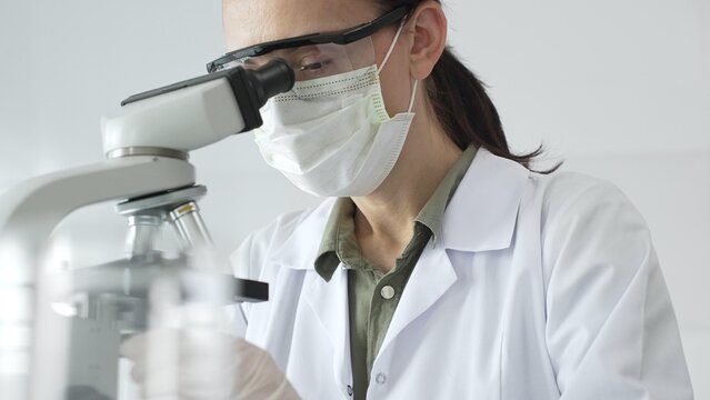 Female scientist examining microscope slide, wearing protective gear while conducting medical research in sterile laboratory environment. Medicine, healthcare and science concept