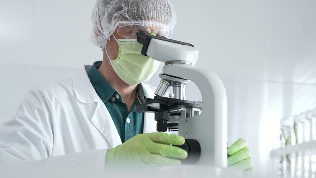 Medical scientist wearing protective mask, cap and green gloves using microscope doing scientific research in laboratory, developing vaccine or medication. Medicine, healthcare and science concept
