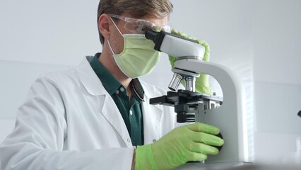 Adult male scientific professional analyzing sample through microscope, wearing green protective gear in sterile laboratory setting. Medicine, healthcare and science concept
