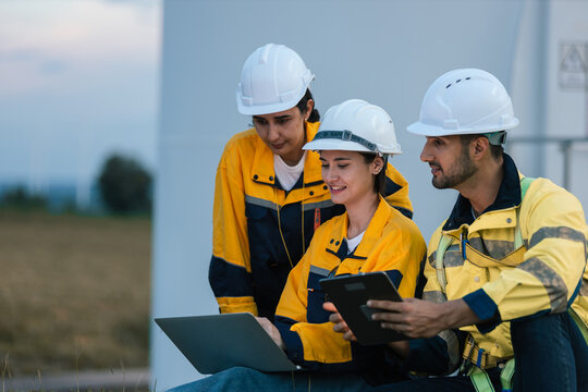 Group of engineers wearing safety helmets and uniforms working together outdoors at wind farm, using laptop and tablet for data analysis, teamwork and sustainable energy project planning. - Powered by Adobe