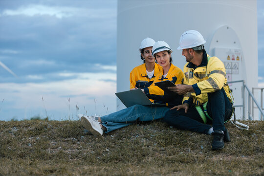 Group of engineers wearing safety helmets and uniforms working together outdoors at wind farm, using laptop and tablet for data analysis, teamwork and sustainable energy project planning.