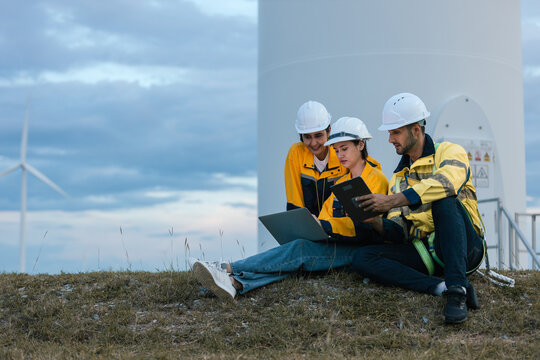 Group of engineers wearing safety helmets and uniforms working together outdoors at wind farm, using laptop and tablet for data analysis, teamwork and sustainable energy project planning.