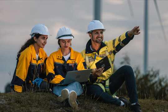 Team of engineers in working at wind farm site during sunset, analyzing renewable energy data using laptop and tablet, collaborating on sustainable power solutions outdoors.