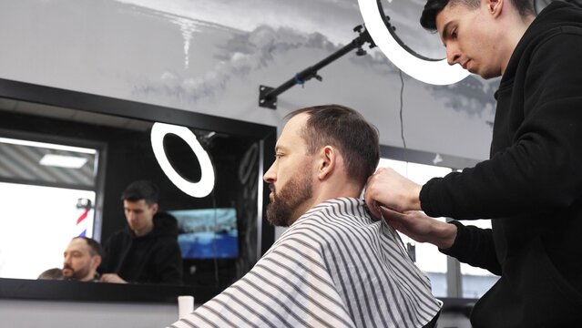 Professional barber removing protective cape from satisfied client, mirror reflecting busy grooming environment with colleague cutting hair in background
