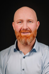 Bearded man with a friendly smile, wearing a light blue shirt, poses against a dark background, showcasing confidence and approachability in a professional setting