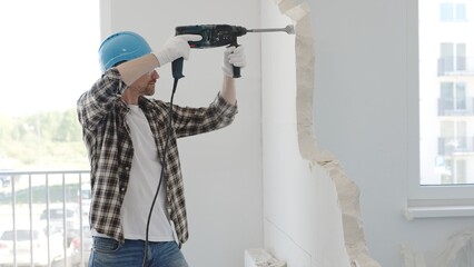 Male construction worker demolishing a wall with a hammer drill in protective gear, creating...
