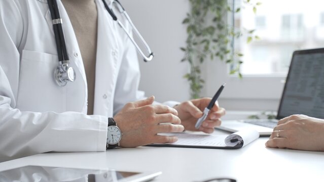 Doctor man is explaining diagnosis to female patient during a medical consultation in a medical office, gesturing with hands over clipboard and white desk. Medicine and health care concept