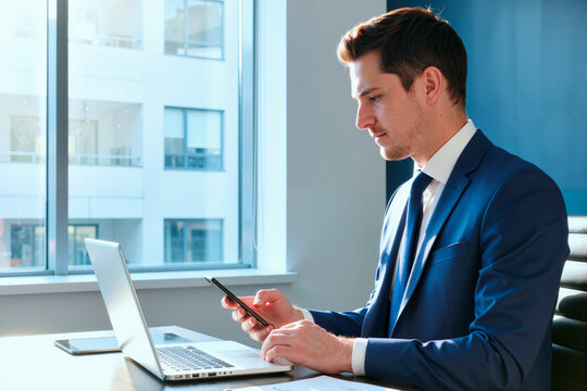 Caucasian young adult man sitting at desk using smartphone and working on laptop in modern office environment, focusing on multitasking and digital communication during business day