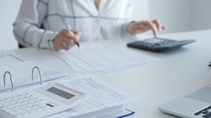 Fototapeta premium White calculator is on financial documents on the office desk, with an accountant taking notes, emphasizing accounting, finance, and tax preparation, close up. Audit and taxes concept