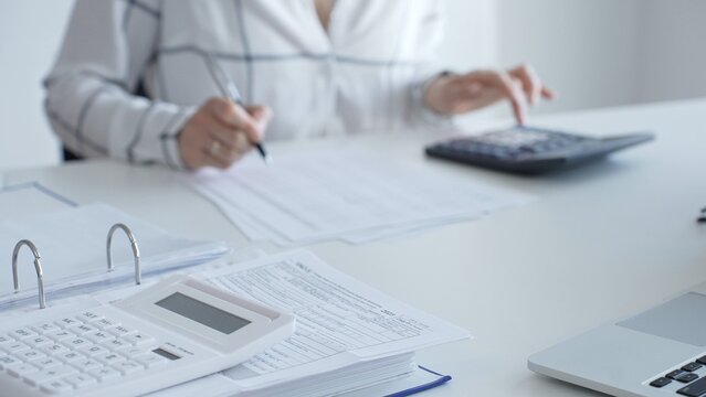 White calculator is on financial documents on the office desk, with an accountant taking notes, emphasizing accounting, finance, and tax preparation, close up. Audit and taxes concept - Powered by Adobe