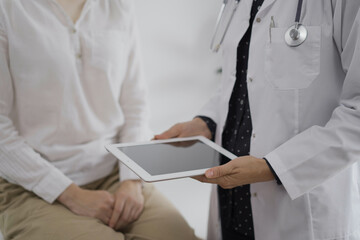 Doctor and a patient discussing something. The female physician, wearing a white medical coat is keeping tablet computer with a medical notes besides a young woman during a consultation in the clinic
