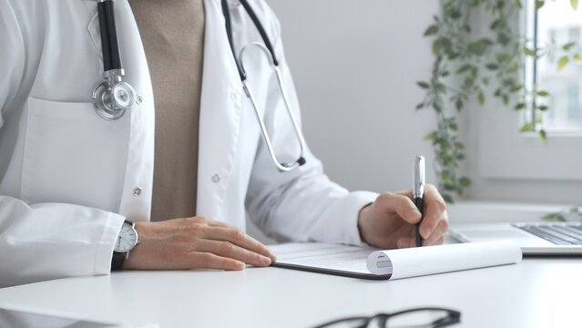 Male doctor working at desk, writing prescription, wearing white lab coat and stethoscope in clinical workspace, close up. Medicine and health care concept - Powered by Adobe