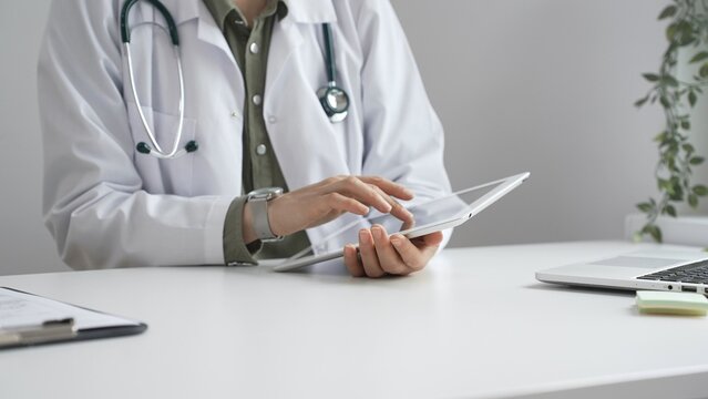 Doctor woman in lab coat and stethoscope using digital tablet at desk in medical office, accessing online patient information. Medicine and health care concept
