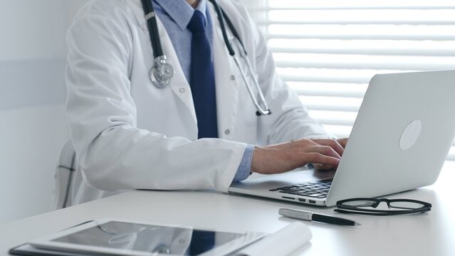 Medical doctor wearing lab coat and stethoscope is sitting at desk and typing on laptop, with eyeglasses and pen nearby, in medical office. Medicine and health care concept