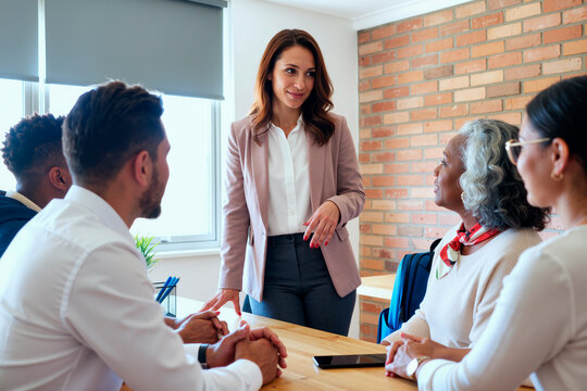 Caucasian young adult woman standing and speaking to diverse group including Black senior woman and two Caucasian young adult men seated at table during business meeting