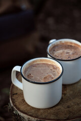 Two white mugs with cocoa in closeup on a tree stump in a forest camp on the background of a hiking backpack