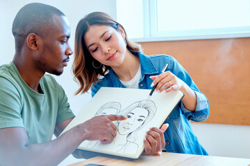 Young adult Black man and young adult Asian woman collaborating while drawing portrait sketches in sketchbook, man pointing at illustration as woman holding pencil and looking at drawing