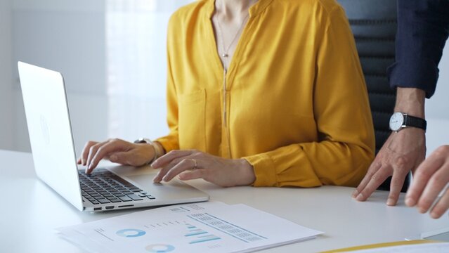 Business partners are collaborating on a project, analyzing financial charts and using a laptop to input data, working together in sunny office. Businesspeople concept