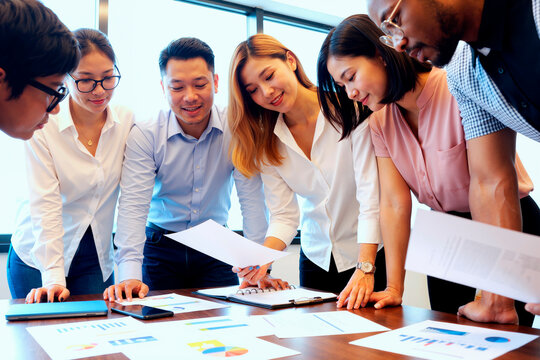 Group of young adult multiethnic men and women collaborating around table, analyzing financial charts and discussing business strategy during office meeting, documents and graphs visible