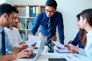 Group of young adult multiethnic colleagues collaborating around table, Asian man standing and handing documents to Caucasian man while others reviewing charts and working on laptop