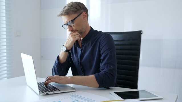 Professional businessman sitting at a sleek desk, working on a laptop and thinking about goals in modern office filled with natural light. Business people concept - Powered by Adobe