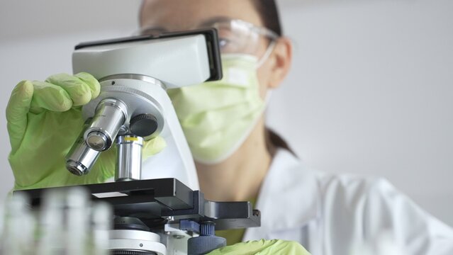 Close up of scientific precision involving green gloved hands adjusting microscope lens, blurred laboratory glassware highlighting medical investigation. Medicine, laboratory and science concept