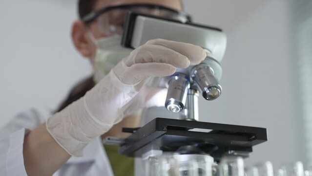 Female scientist wearing white protective gear analyzing test samples under microscope in contemporary research environment, close up of white gloved hand. Medicine and science concept