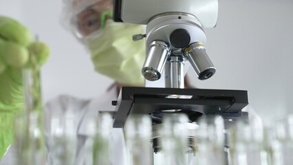 Scientist wearing green gloves and a face mask selecting a test tube with transparent liquid from a rack near a microscope in a lab. Medicine, healthcare and science concept