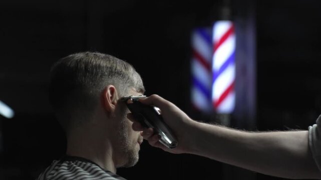 Skilled barber trimming client's hair using electric clipper, vintage barbershop interior with blurred pole, professional grooming service