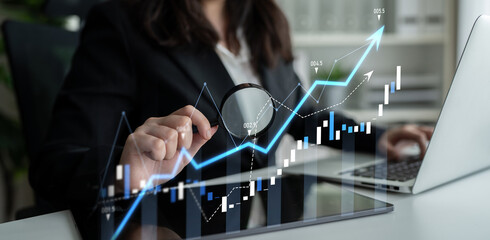 A businesswoman analyzes financial graphs using a laptop and magnifying glass, reflecting a modern approach to economic trends and market research in a sleek office environment. Scalp