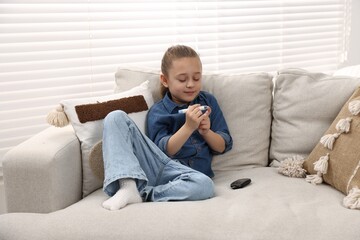 Diabetes. Little girl checking blood sugar level with lancet pen on sofa indoors