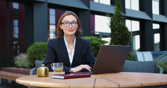 Close up portrait smiling face business woman looking at camera wearing stylish glasses sipping coffee and typing on laptop posing outdoors during lunch break of the cafe. Female manager employer