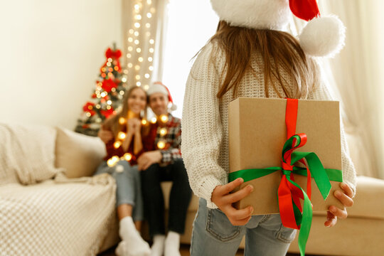 Little girl in Santa hat holding a Christmas present with red and green ribbon behind her back, parents sitting on the sofa with lights and tree in the background, family love and festive joy