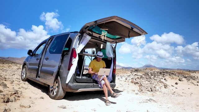 Mature male digital nomad working as a freelancer on his laptop, sitting on the trunk of his camper minivan in a beautiful, sunny and wild volcanic landscape with a blue sky background