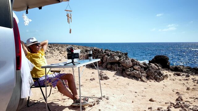 Content creator working on his blog at a desk set up behind his minivan on a sunny volcanic beach, enjoying the freedom of a nomadic lifestyle and remote work in a scenic ocean setting
