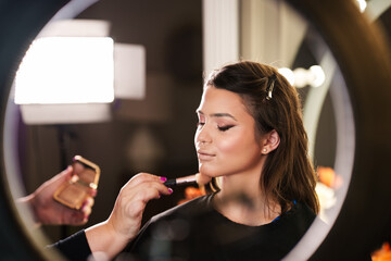 Professional beautician applying makeup on beautiful young woman's face. Professional make up beauty studio work with gorgeous female model.