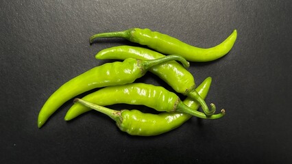 Close-up photo of fresh green chili peppers arranged on a dark surface. The vibrant color and glossy texture highlight the freshness of the peppers, perfect for use in food, spice, and cooking.