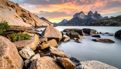 A serene landscape features jagged peaks and a calm lake at sunset. Rocks, vegetation, and an orange-streaked sky complete the scene