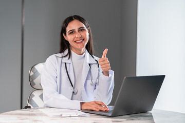 Confident female doctor smiling and showing thumbs up while working with a laptop in a modern clinic office, representing successful healthcare service, digital consultation, and professionalism.