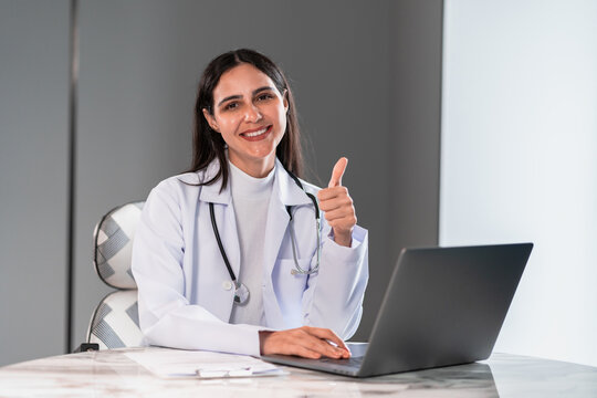 Confident female doctor smiling and showing thumbs up while working with a laptop in a modern clinic office, representing successful healthcare service, digital consultation, and professionalism. - Powered by Adobe