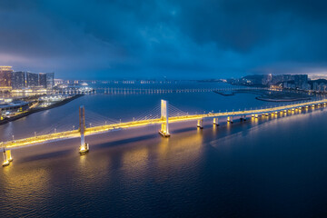 Aerial night view of the beautifully illuminated cable-stayed bridge spanning the sea under a dramatic cloudy blue sky in Macau.