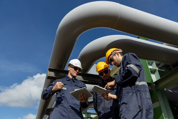 Engineer wearing safety gear using digital tablets at an industrial facility, discussing data and operations under large pipelines, symbolizing technology integration in modern engineering.